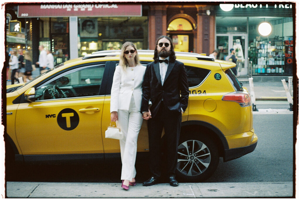 Film wedding photographer documents stylish couple in white suit and tuxedo standing on Brooklyn Bridge during foggy New York City elopement.