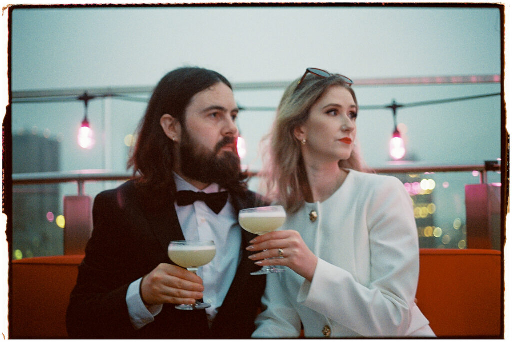 NYC elopement photographer captures couple in black tie and white suit drinking martinis on a rooftop at sunset, shot on film.