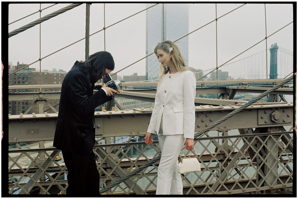 Film wedding photographer documents stylish couple in white suit and tuxedo standing on Brooklyn Bridge during foggy New York City elopement.