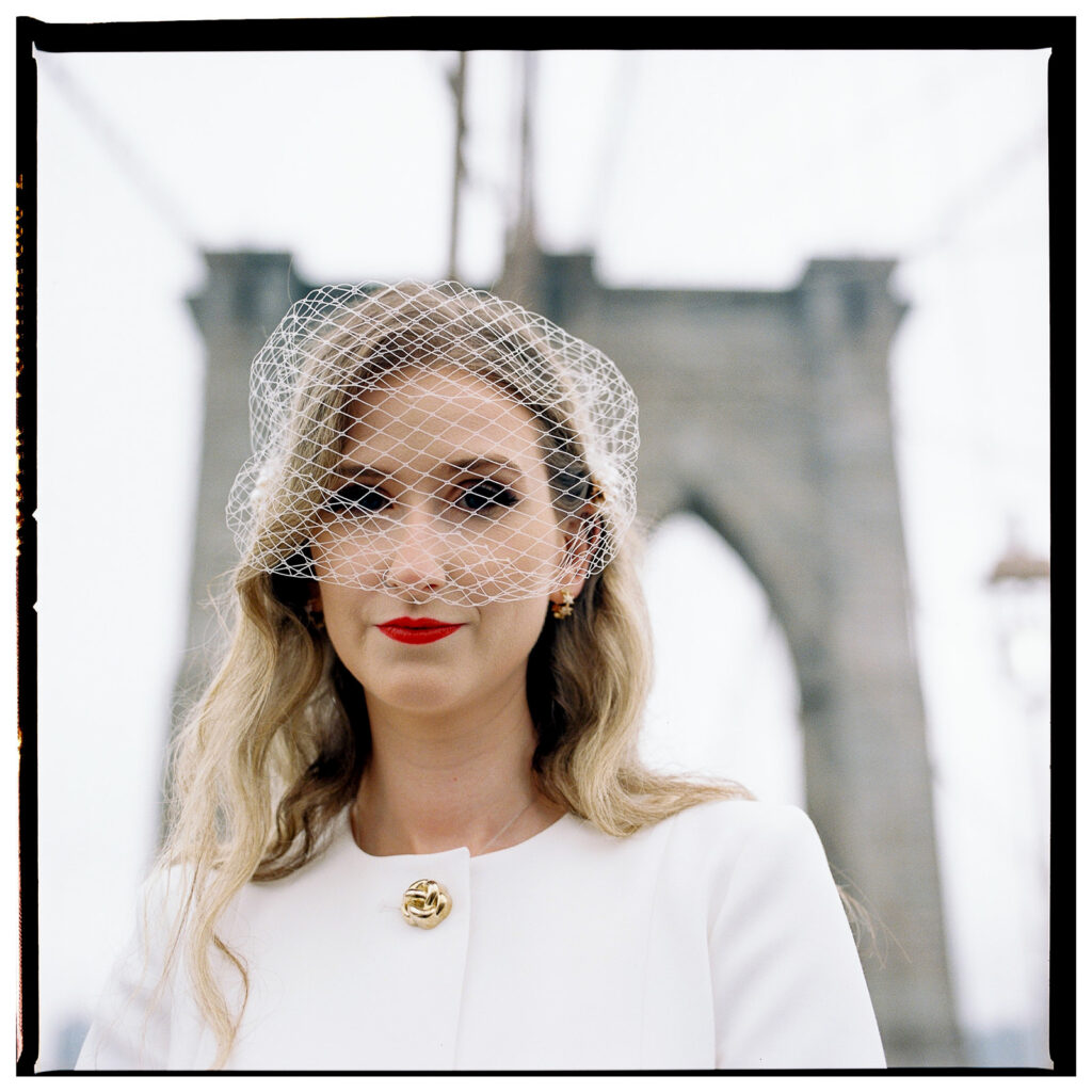 Film wedding photographer documents stylish couple in white suit and tuxedo standing on Brooklyn Bridge during foggy New York City elopement.