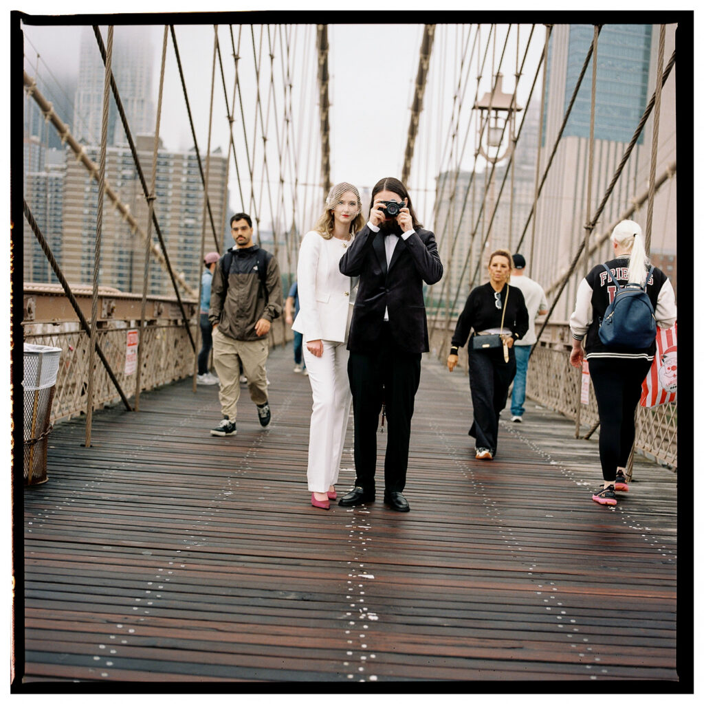 Film wedding photographer documents stylish couple in white suit and tuxedo standing on Brooklyn Bridge during foggy New York City elopement.