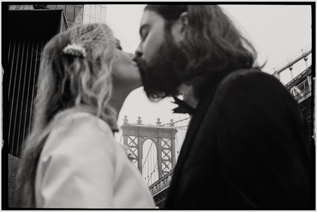 Film wedding photographer documents stylish couple in white suit and tuxedo standing on Brooklyn Bridge during foggy New York City elopement.