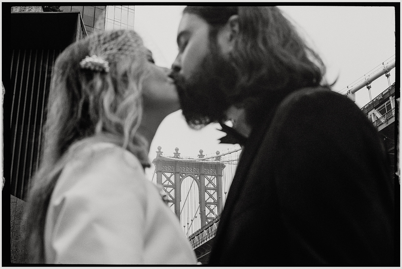 Film wedding photographer documents stylish couple in white suit and tuxedo standing on Brooklyn Bridge during foggy New York City elopement.
